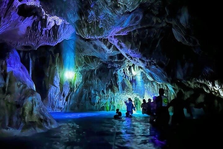 The cave interior lit in blue and violet, visitors standing in the water